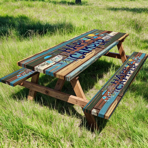 Outdoor wood picnic table and benches with 'Happy Camper' design, placed on grass, surrounded by trees in a sunny setting.