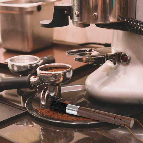 Takavu coffee grinder cleaning brush with wooden handle and black boar bristles beside a stainless espresso machine.