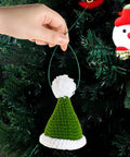 Hand holding green and white knit hat ornament hanging on a Christmas tree