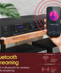 Bluetooth 5.1 Home Theater And Karaoke Amplifier That Delivers Party Ready Big Room Sound With Five Channels And Subwoofer Output Two Mic Inputs With Echo USB A Plus SD MP3 Player RCA In PDA8BU on shelf with phone showing music streaming.