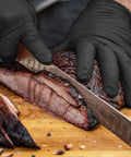 Person slicing meat while wearing Grill Ready Disposable BBQ Gloves Kit With 50 Nitrile Gloves, Powder Free, Latex Free, Food Safe.