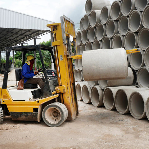 Forklift extensions on a yellow forklift lifting a concrete pipe amid stacked concrete pipes