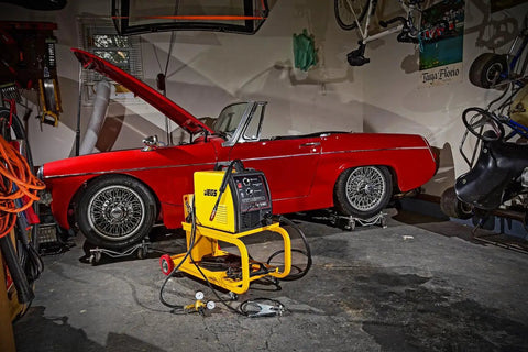 HEAVY DUTY YELLOW POWDER COATED STEEL WELDING CART WITH SMOOTH ROLLING CASTERS AND SLANTED WORK SURFACE BUILT TO CRUSH THE GRIND AND HAUL YOUR GEAR FAST in a garage beside a red convertible car.