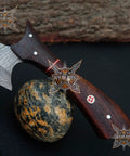 Beast Damascus Steel Skinner Knife with wood handle, showing blade detail resting on a rock, part of outdoor hunting gear.