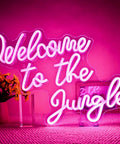 Welcome To The Jungle Neon Sign Pink LED Word Neon Light USB Powered Letter Light in brightly lit room with flowers.
