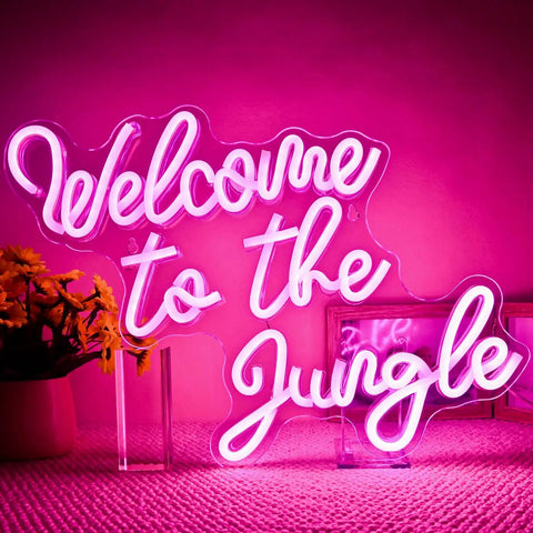 Welcome To The Jungle Neon Sign Pink LED Word Neon Light USB Powered Letter Light in brightly lit room with flowers.