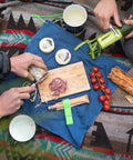 Picnic scene with Nomad Camping Kitchen Utensil Kit, No 12 serrated knife slicing sausage, No 6 peeler peeling cucumber.