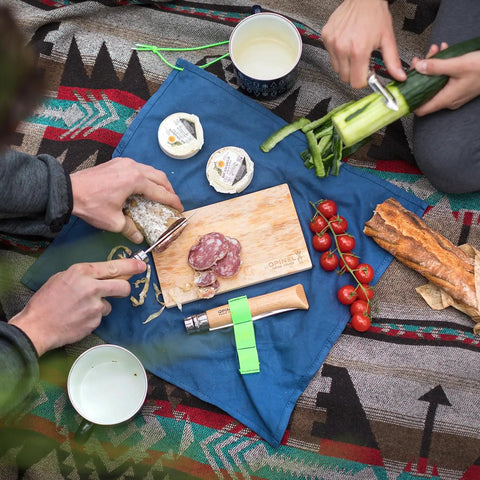 Picnic scene with Nomad Camping Kitchen Utensil Kit, No 12 serrated knife slicing sausage, No 6 peeler peeling cucumber.
