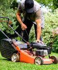 Person starting a red lawn mower on a grassy lawn, wearing a cap and vest, surrounded by trees and bushes.
