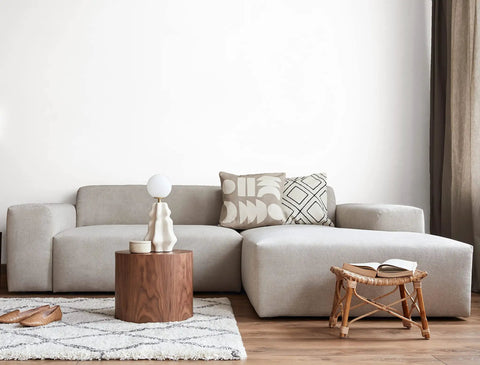 Modern living room with a beige sectional sofa, wooden coffee table, decorative pillows, and a small stool on a patterned rug.