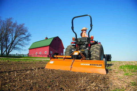 Farmer using rotary tiller on field with red barn in background