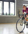 Cyclist using an indoor trainer with Front Wheel Block For Indoor Bike Trainers Level Your Ride And Boost Stability 11x5x4 Inch Riser Keeps Your Front Wheel Aligned With The Trainer For A Realistic Ride Easy Setup And Durable Support.