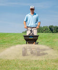 Spyker All Season Spreader Kit Ergo Pro 50 Pound Broadcast Spreader with 25 Pound Bag in use on lawn by person pushing.