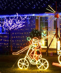 Festive yard with Christmas lights display featuring an illuminated snowman on a bicycle and colorful tree lights in the background.