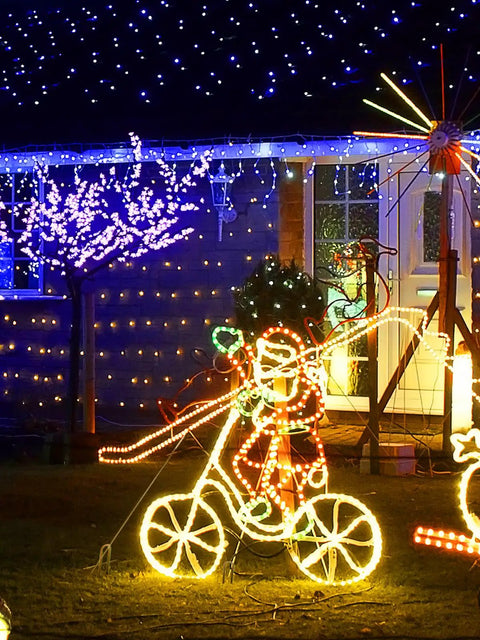 Festive yard with Christmas lights display featuring an illuminated snowman on a bicycle and colorful tree lights in the background.
