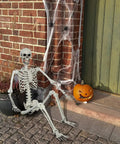 5.4Ft Life Size Halloween Skeleton With Movable Joints sitting by a brick wall with cobwebs, a pumpkin, and a door.