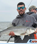 Angler holding a striped bass fish on a boat with fishing gear visible, wearing sunglasses and a hoodie.