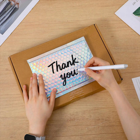 Person writing 'Thank you' on 60 Pack Holographic Bubble Mailers Dope Waterproof Self Seal Foil Glitter Glamour Shipping Envelopes In 3 Sizes For Mailing Jewelry, Makeup, And Small Business Packaging Needs