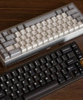 Two compact 65 Percent Wireless Mechanical Keyboards in gray and black with CNC aluminum bodies on a wooden desk.