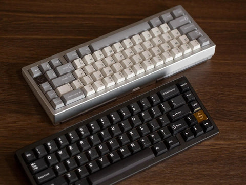 Two compact 65 Percent Wireless Mechanical Keyboards in gray and black with CNC aluminum bodies on a wooden desk.
