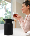 Woman enjoying a drink beside a 50 Oz Stainless Steel Thermal Coffee Carafe Insulated Vacuum Double Walled Beverage Dispenser With Tea Infuser And Strainer For Hot Or Cold Drinks Keeps Your Brew Hot Or Cold For Hours