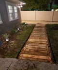 Wooden walkway in a backyard secured with plastic tarp stakes at night, surrounded by garden lighting and bordered by lawn edging.
