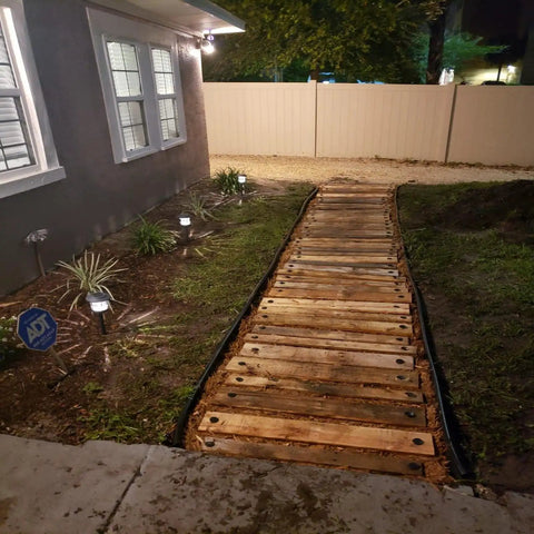 Wooden walkway in a backyard secured with plastic tarp stakes at night, surrounded by garden lighting and bordered by lawn edging.