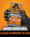 Close-up of a gorilla hand holding a pack of crystal clear adhesive dots, promotional text about no mess and instant hold visible.