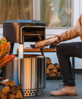 Bonfire Pi Fire Pizza Oven Accessory For Fire Pit With Cordierite Pizza Stone And Carry Case Wood Burning Assembly Stainless Steel Build Elevate Pizza Nights Grillin On The Pit in use with wood being added to fire pit.