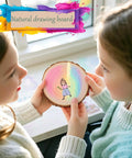 Two children holding a wood slice with a colorful drawing, labeled 'Natural drawing board', near a bright window.
