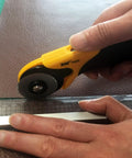 Hands using 45mm Rotary Cutter And Precision Knife Set For Art Quilting And Crafting With 12 Bonus Blades Pro Grade Ergonomic Aluminum Handle Anti Slip Grip Safe Retracting Blade For Precise Clean Cuts on fabric with ruler.