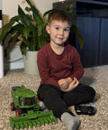 Child playing with a Remote Control Combine Harvester Tractor Toy With Moving Corn Head RC Farm Tractor For Kids 3 Plus Light Sound Spray Effects One Twenty Fourth Scale Farm Toy Totally Perfect Christmas Birthday Gift on a carpeted floor with a plant in the background.
