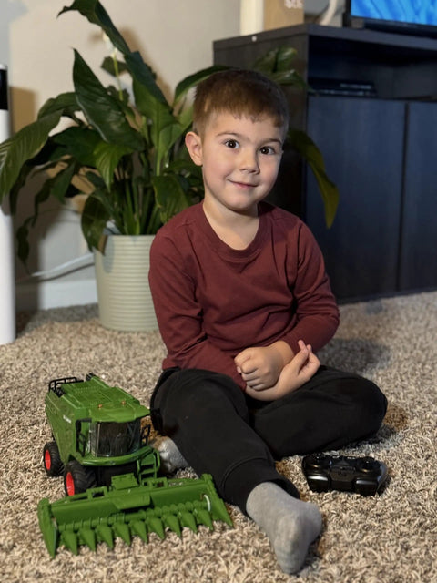 Child playing with a Remote Control Combine Harvester Tractor Toy With Moving Corn Head RC Farm Tractor For Kids 3 Plus Light Sound Spray Effects One Twenty Fourth Scale Farm Toy Totally Perfect Christmas Birthday Gift on a carpeted floor with a plant in the background.