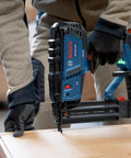 Gutsy 18 Volt Brushless Cordless Brad Nailer Kicks Out 2,000 Nails Per Charge With One Thumb Safety Trigger Zero Hassle Jam Clearance And Built In Dual Lamps For Low Light Jobs No Battery Or Charger Included, being used to nail wood.