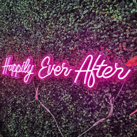 Pink Happily Ever After Neon Sign Wall Art Hangin Neon Light For Party Vibes Wedding Hashtag Decor Kid Bedroom Bar Home 12V DC Power Adapter Included For Easy Setup glowing against green leaves.