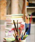 A variety of paintbrushes and pencils in a holder on a workspace, with art supplies in the background, showcasing creativity.