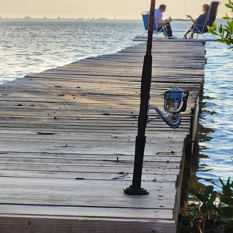 Fishing rod resting on a wooden dock over water with two people sitting in chairs in the background