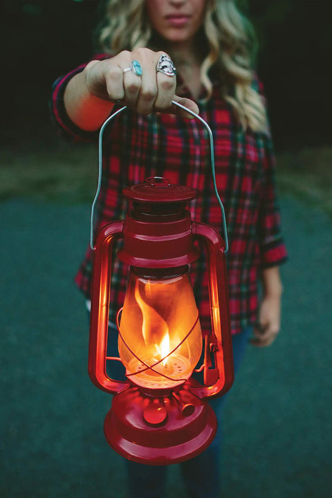 Person holding 12 Inch Hurricane Lantern With Durable Red Finish For Camping And Emergencies glowing brightly in the evening.