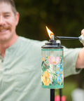Person lighting a Wildflower Glass Outdoor Torches 2-Pack, 65-Inch Easy Install Patio Backyard Lighting for Stylish Lawn Ambience.