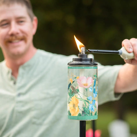 Person lighting a Wildflower Glass Outdoor Torches 2-Pack, 65-Inch Easy Install Patio Backyard Lighting for Stylish Lawn Ambience.