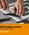 Fiskars self-healing cutting mat with grid on gray surface; hands slice black foam shapes with a craft knife.