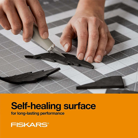 Fiskars self-healing cutting mat with grid on gray surface; hands slice black foam shapes with a craft knife.