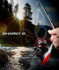 Angler holding Sharky III 3000 Spinning Fishing Reel for Saltwater and Freshwater over scenic river at sunset, showing graphite build