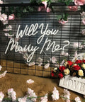 Will You Marry Me Neon Light Sign For Wedding Decor And Proposal Night Light Up Letters For Get Hitched Bedroom Wall Art Romantic Gift For Women 23 By 14 Inches LED Accent For Home And Party glowing amidst floral backdrop.