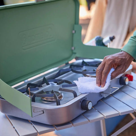 Person cleaning burners of Three In One Outdoor Camp Stove With Removable Cast Iron Grill And Griddle 24,000 BTUs on metal table