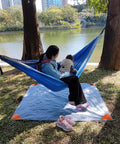 A person and child sitting in a hammock over a Pocket Picnic Blanket For Grab And Go Outdoor Fun That Folds Small And Lightweight Sandproof 55 By 60 Inch Beach Mat Water Resistant Quick Dry Washable Ultra Portable Tarp For Camping Picnics Festivals Parks And Outdoor Recreation by a lakeside.