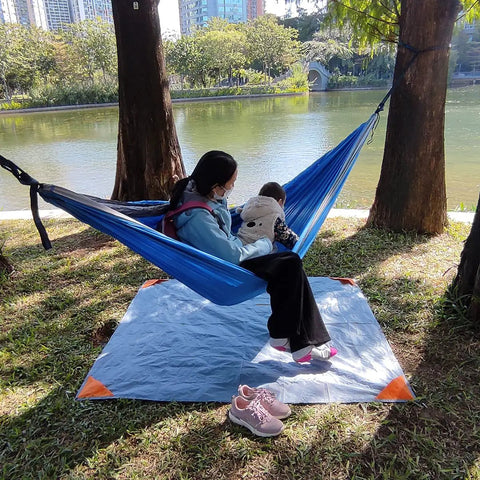 A person and child sitting in a hammock over a Pocket Picnic Blanket For Grab And Go Outdoor Fun That Folds Small And Lightweight Sandproof 55 By 60 Inch Beach Mat Water Resistant Quick Dry Washable Ultra Portable Tarp For Camping Picnics Festivals Parks And Outdoor Recreation by a lakeside.