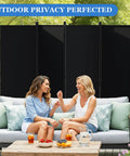 Six Foot Folding Privacy Screen With Lockable Wheels, 4-Panel Room Divider in black used outdoors, behind two women on a sofa