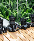 Various Canon DSLR cameras with 58mm Tulip Flower Lens Hoods on a wooden table, surrounded by greenery.