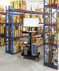 Heavy Lifter Mobile Workstation Cart With No Sweat Rolling Casters And Fully Adjustable Gas Spring Monitor Stand Plus Secure CPU Tray For Seamless Standing Desk Transitions In Matte Black in a warehouse with boxes.